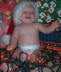 A baby in diapers laughs while surrounded by Christmas lights and wearing a Santa hat