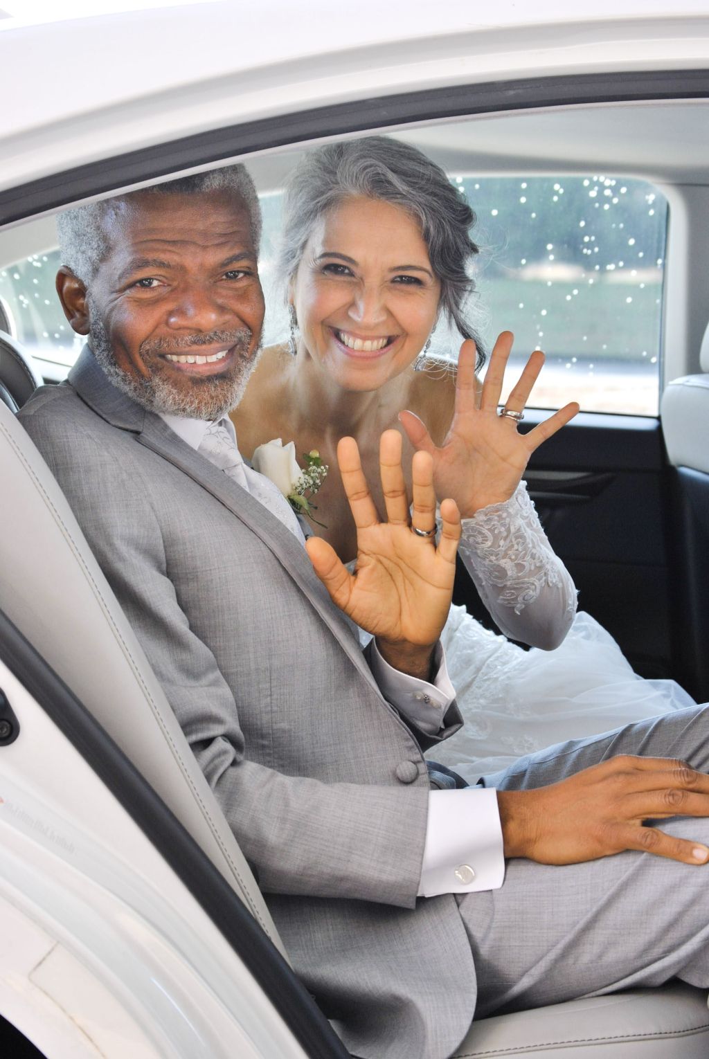 A beautiful Argentinian Christian woman beams with joys while seated in a car next to her new American husband, as both show wave and show off their new wedding rings