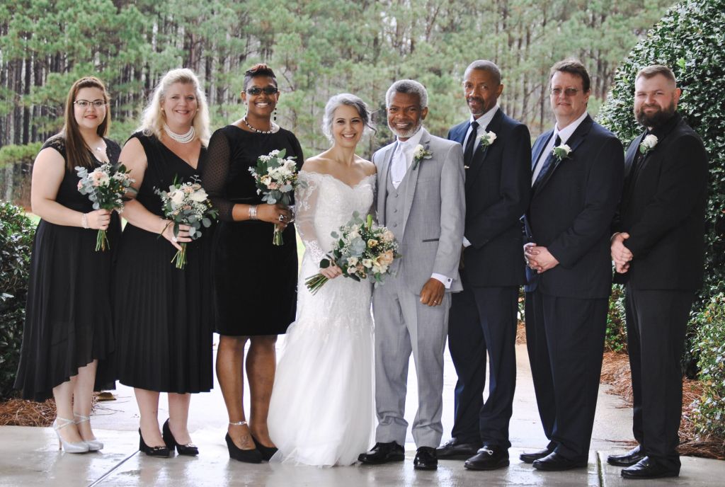 A middle aged Christian couple surrounded by their wedding party pose on the edge of a forest