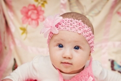 A baby girl with a pink headband and flower stares curiously at the camera