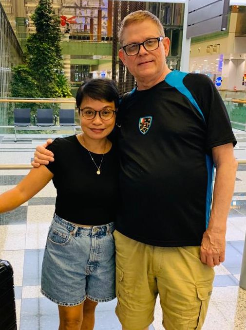 An Asian woman smiles at a mall as a White man pulls her in close for a hug