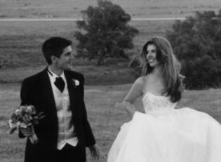 A bride tries running in a field while holding her dress. Husband holds her flowers and laughs
