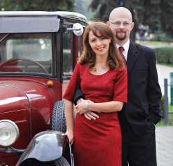 A beautiful European woman in a red dress leans into a man standing next to a vintage car