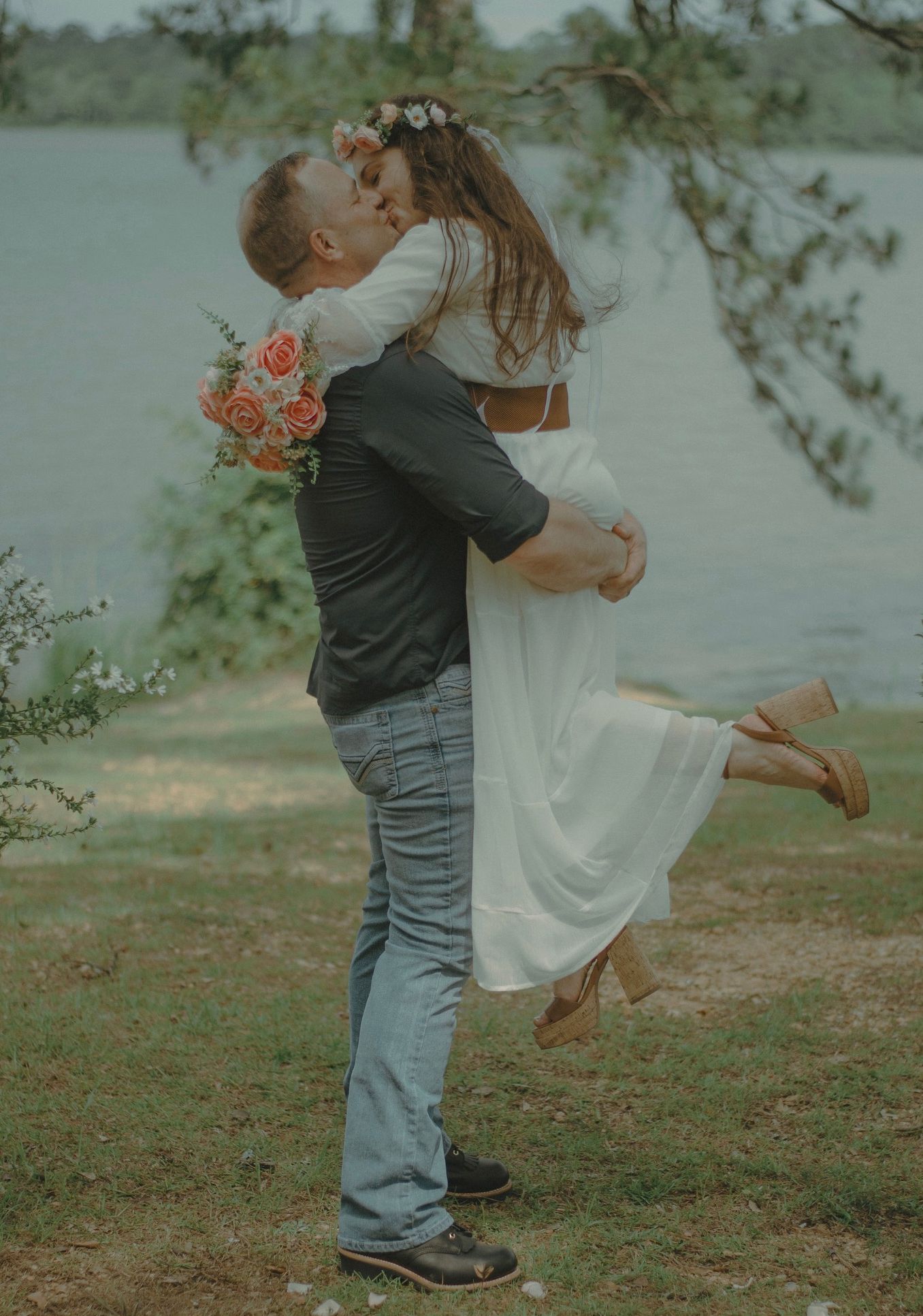An excited bride leaps into her new husband's arms for a kiss. Her husband holds her above the ground as they kiss in front of a beautiful lake