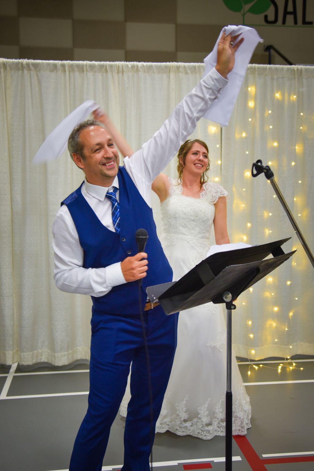 A groom holds a microphone while he and his bride encourage their audience to celebrate their union