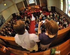 A Christian couple stand in a church and take a picture while in the background their friends marry