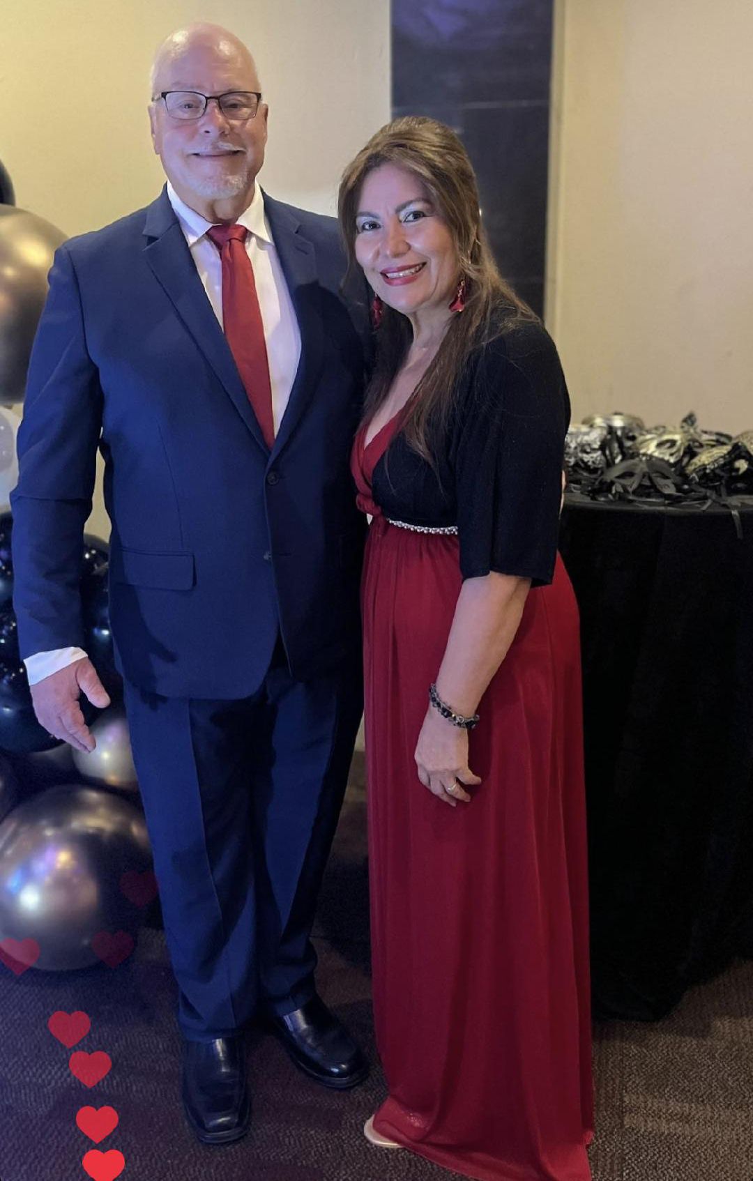 A woman in a flowing red dress and medium length hair smiles as a man in a blue suit stands next to her