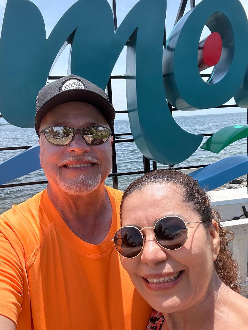A man in an orange t-shirt, wearing sunglasses and a baseball hat smiles at the camera as a woman in glasses leans in. The ocean is seen in the background