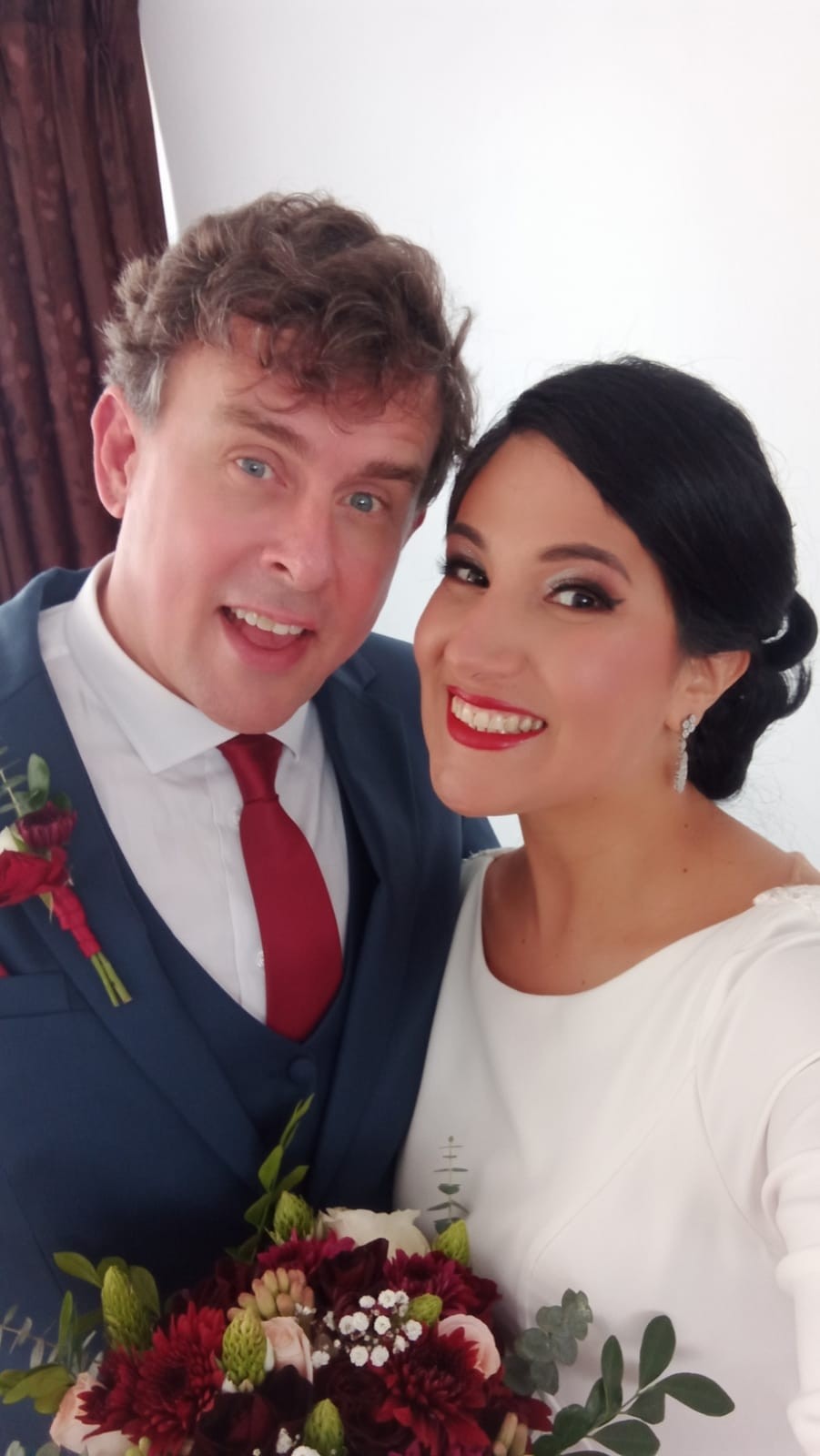 A White man looks overjoyed standing next to his new Latina bride, who smiles in a white dress while holding a bouquet of flowers