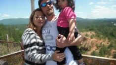 A dad looks lovingly at his daughter while his wife hugs him closely in this outdoor family shot