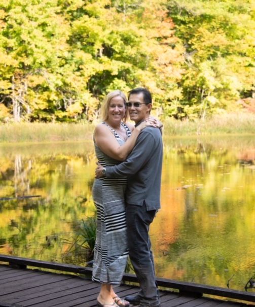 A very happy couple hug on a dock overlooking water and Autumn colours