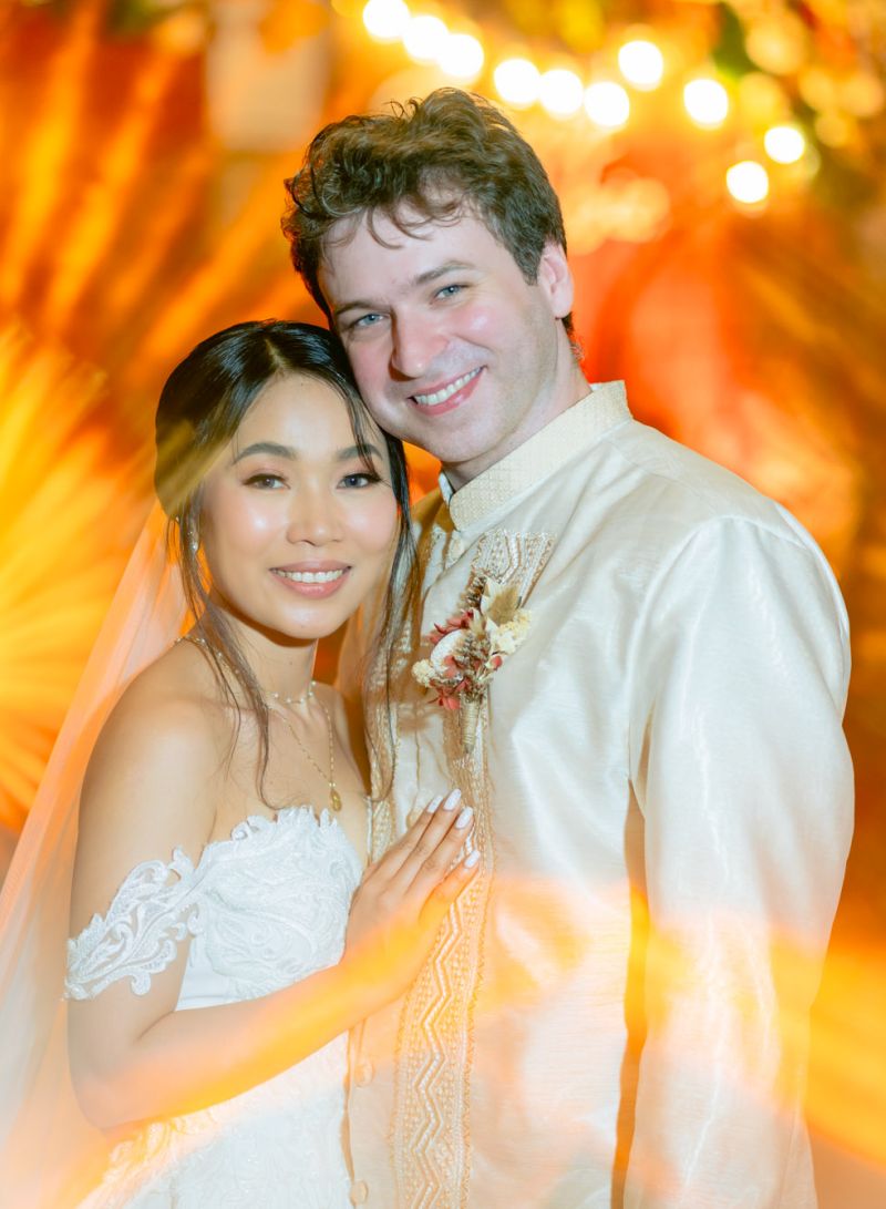 A man with blue eyes and wearing a boutonniere leans into his beautiful Asian bride as she places her hand affectionately on him and smiles at the camera