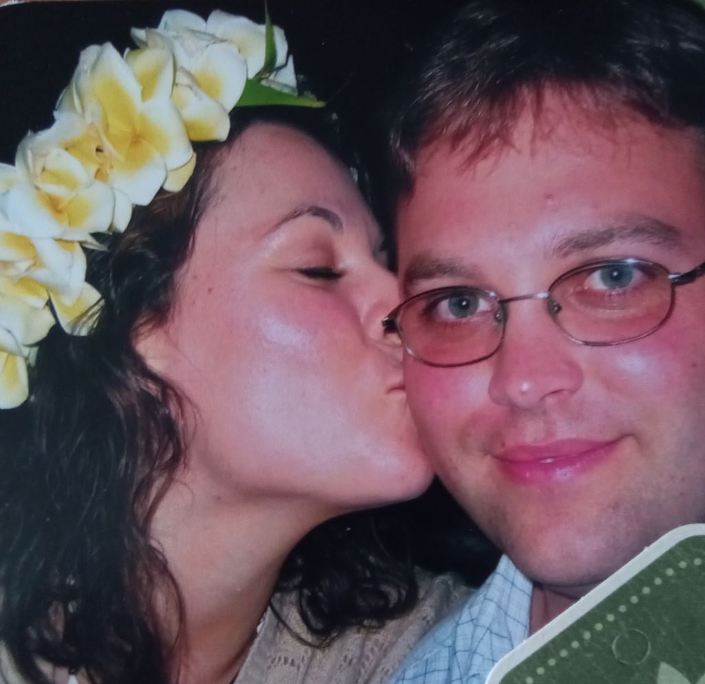 A Christian woman lovingly kisses her smiling husband on the cheek, while wearing a Hawaiian lei