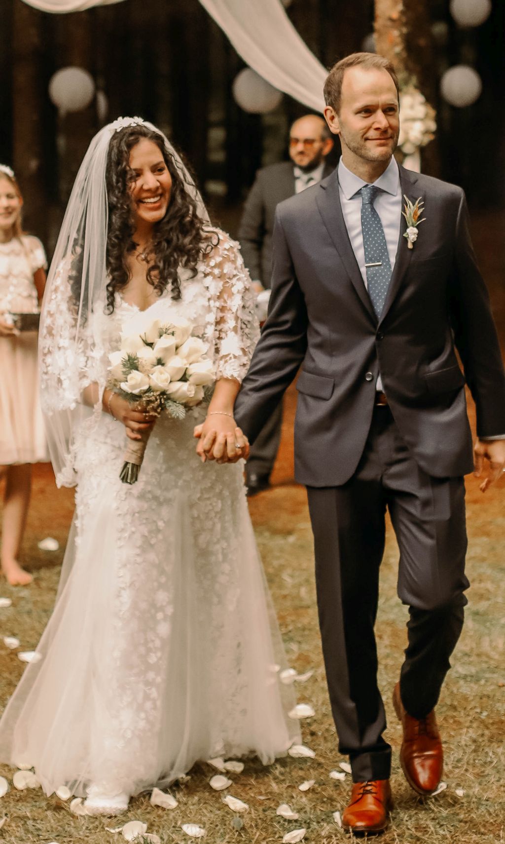 A Christian bride in a beautiful white wedding dress smiles with sheer joy as she holds a bouquet of flowers in one hand, and her new husband's in the other