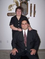A proud man is seated and smiling as his wife stands behind and hugs him
