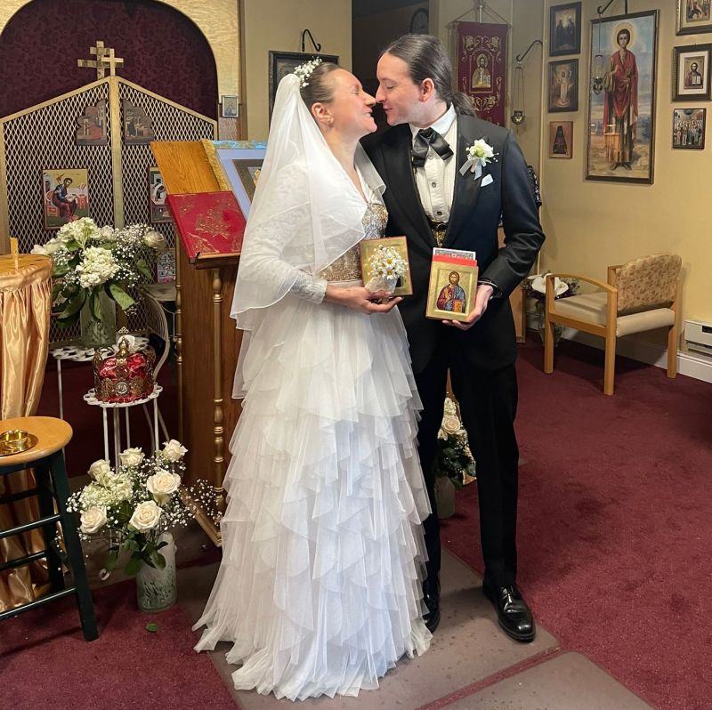 A new bride in white dress kisses her tuxedo husband while they each hold photos of saints