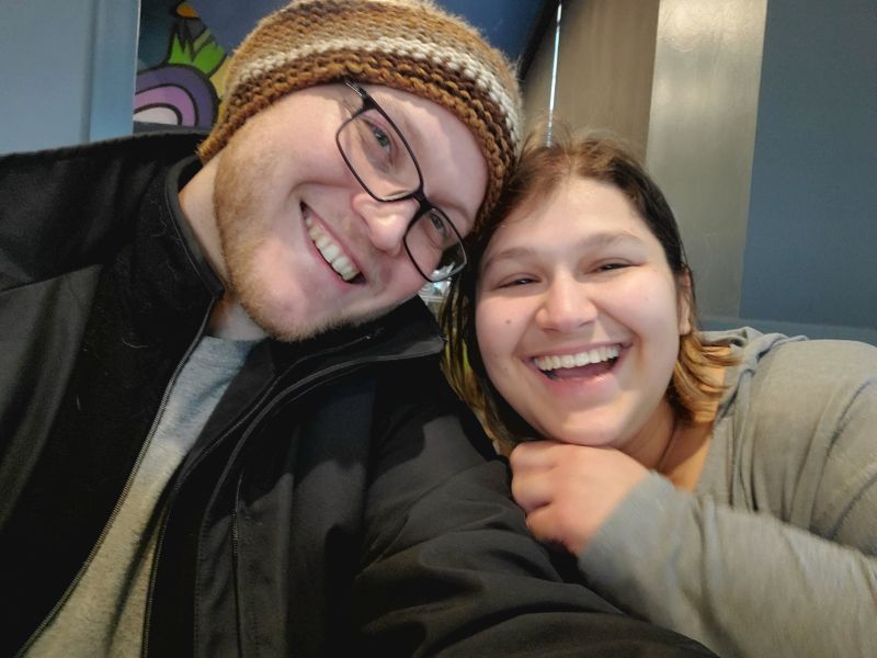 A Christian single man wearing glasses and toque laughs next to a woman who smiles with him for a selfie