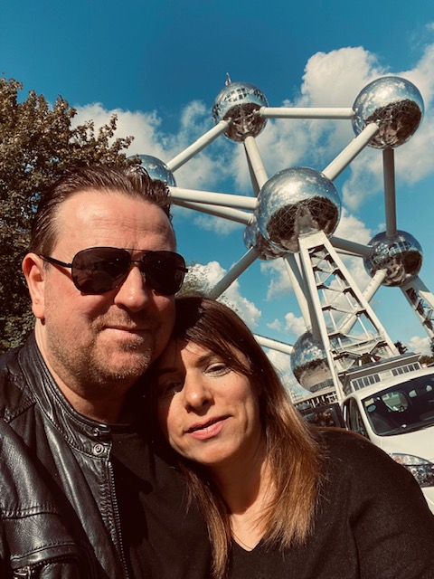 A man in sunglasses smiles while a woman leans her head on his shoulder, outside the Atomium in Belgium