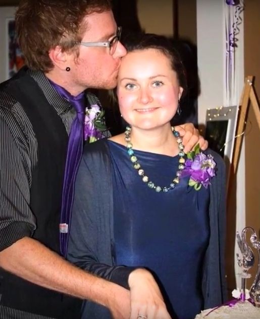 Man affectionately kisses top of woman's head as she beams with joy