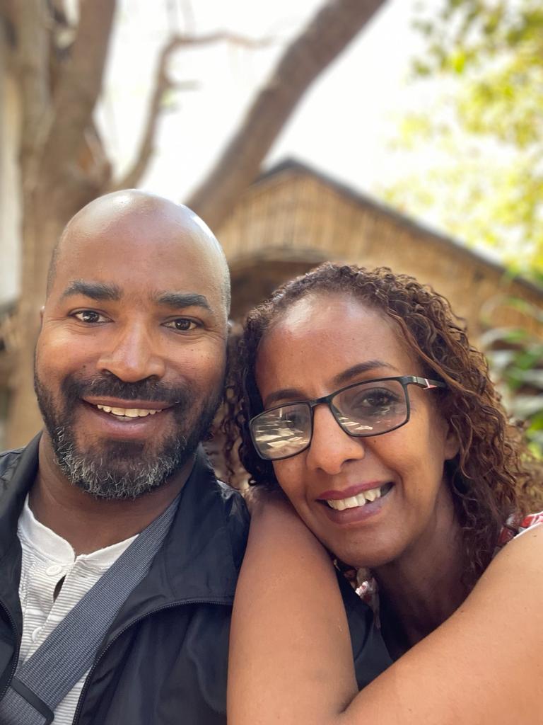 A relieved woman wearing glasses leans on a handsome man who looks pleased to be with her, as they stand outdoors in Ethiopia.
