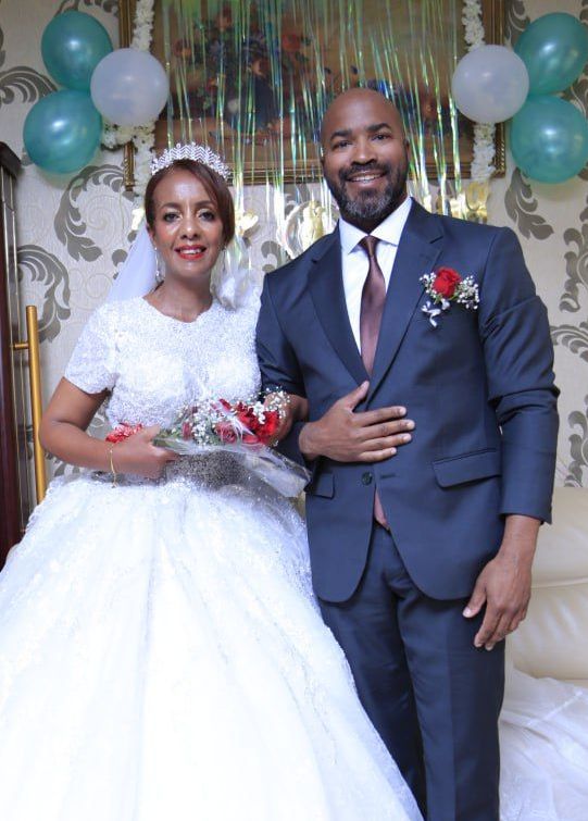 A beautiful African Christian bride and her American husband stand proudly after marrying. Blue and white celebration balloons in the background