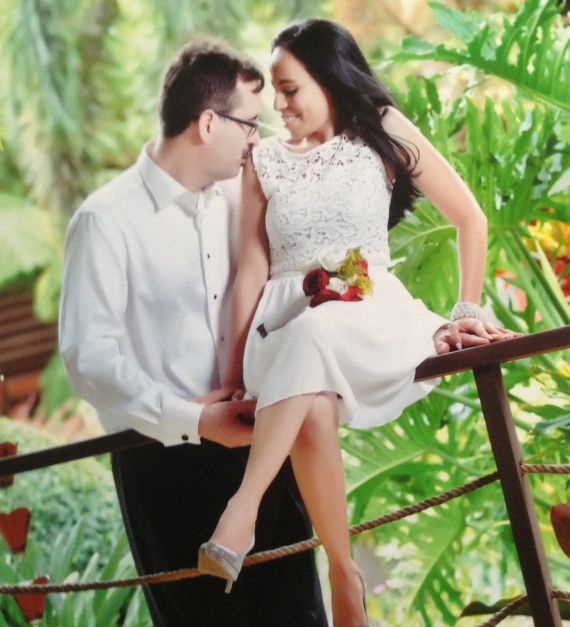 Beautiful bride sits on a fence while husband supports her and kisses her shoulder