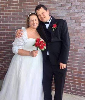 A joyful bride in beautiful white wedding dress and red bouquet is hugged by her husband in tuxedo