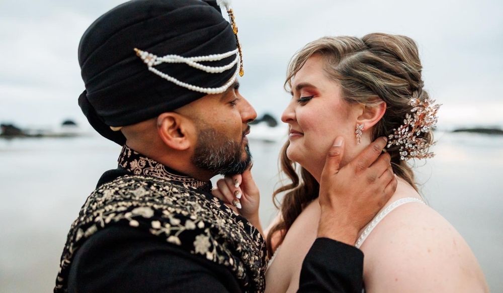 A groom in traditional Indian wedding costume leans in to kiss his American bride